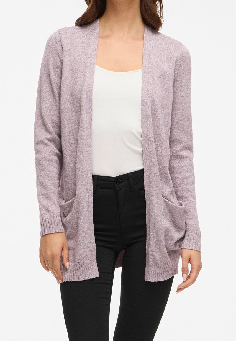 Femme portant un cardigan violet pâle ouvert sur le devant avec des poches, un haut blanc et un pantalon noir, debout devant un fond blanc.