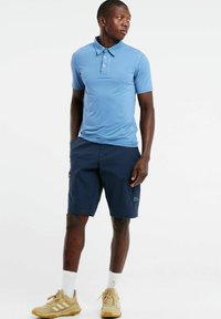 Šviesiai mėlynas polo marškinėlis su mygtukų apykakle, derinamas su tamsiai mėlynomis šortais, turinčiais šoninę kišenę. Dėvima su smėlio spalvos sportiniais batais ir baltais kojiniais.