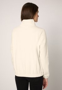 Jasno kremowa kurtka z polaru z wysokim kołnierzem, elastycznym pasem i długimi rękawami. Gładka faktura, minimalistyczny design, brak widocznych elementów metalowych.