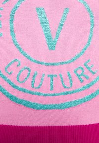 Tissu tricoté avec un logo « V Couture » bleu sarcelle sur un fond rose clair et une bordure rose foncé en bas.