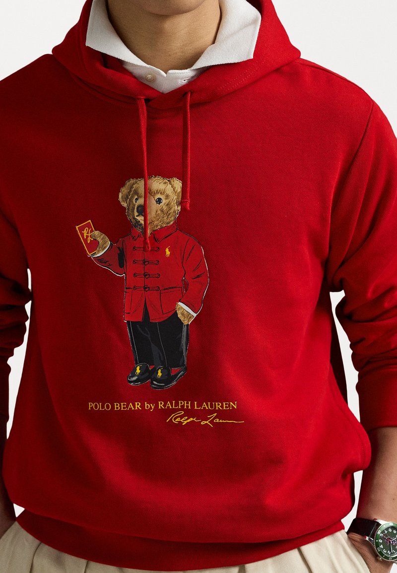 Czerwona bluza z kapturem z haftowanym niedźwiedziem w czerwonym płaszczu trzymającym kartę. Tekst brzmi "POLO BEAR by RALPH LAUREN" w kolorze żółtym.