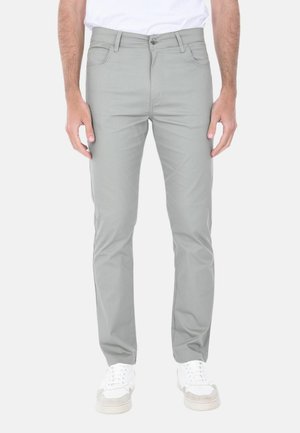 Uomo che indossa pantaloni slim fit grigio chiaro e sneakers bianche, in piedi con le braccia rilassate lungo i fianchi su uno sfondo semplice.