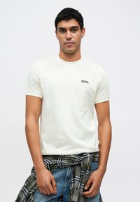 Σύντομη μπλούζα με κοντά μανίκια σε ελαφρύ κρεμ απόχρωση με στρογγυλή λαιμόκοψη, διακοσμημένη με ένα μικρό μαύρο λογότυπο στο αριστερό στήθος. Φορεμένη με πράσινο καρό πουκάμισο δεμένο στη μέση.