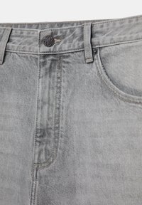 Gros plan sur un jean en denim gris clair montrant le bouton, la ceinture, les passants de ceinture, le haut de la poche avant et les détails de couture.