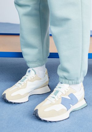 Personne portant un pantalon de survêtement bleu clair et des baskets blanches et beiges avec des touches de bleu, debout sur un tapis bleu près d’un banc en bois.
