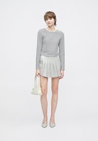 Koszula w paski z długim rękawem i plisowana szara mini spódniczka. Modelka trzyma małą białą torebkę i nosi jasne buty na ostro zakończonych noskach.