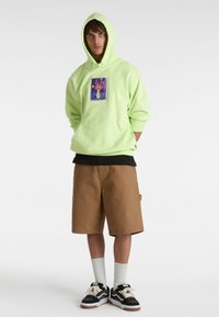 Un sweat à capuche vert citron avec un motif floral, un t-shirt noir en dessous, un short marron, et des baskets Vans noir et blanc avec des chaussettes blanches.