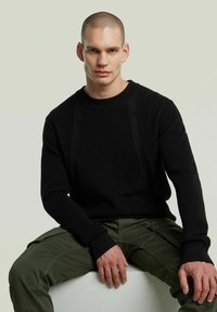 Maglione nero a maglia con collo a giro, caratterizzato da una texture a coste e maniche lunghe. Abbinato a pantaloni cargo verdi. Design minimalista.