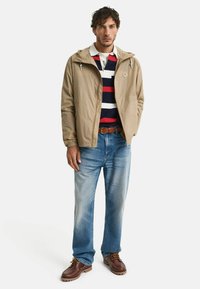 Giacca windbreaker beige chiaro con cappuccio, indossata sopra una polo a righe blu navy, abbinata a jeans blu sbiaditi e stivali marroni con lacci.