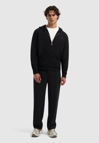 Man met krullend haar in een zwarte zip-up hoodie, zwarte broek, wit T-shirt en witte sneakers, staand tegen een effen lichte achtergrond.