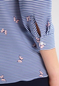 Modrá a bílá pruhovaná blůza s jemným vzorem růžových motýlů, s detaily manžet na knoflíky a mírným rozparkem na rukávech.