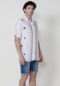 Camisa blanca de manga corta con bordado floral negro en el frente y el cuello. Combinada con pantalones cortos de mezclilla azul, doblados en el dobladillo.