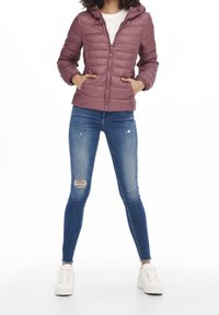 Veste matelassée rose avec capuche, fermeture éclair à l'avant et poches latérales. Portée avec un jean skinny bleu déchiré et des baskets blanches.