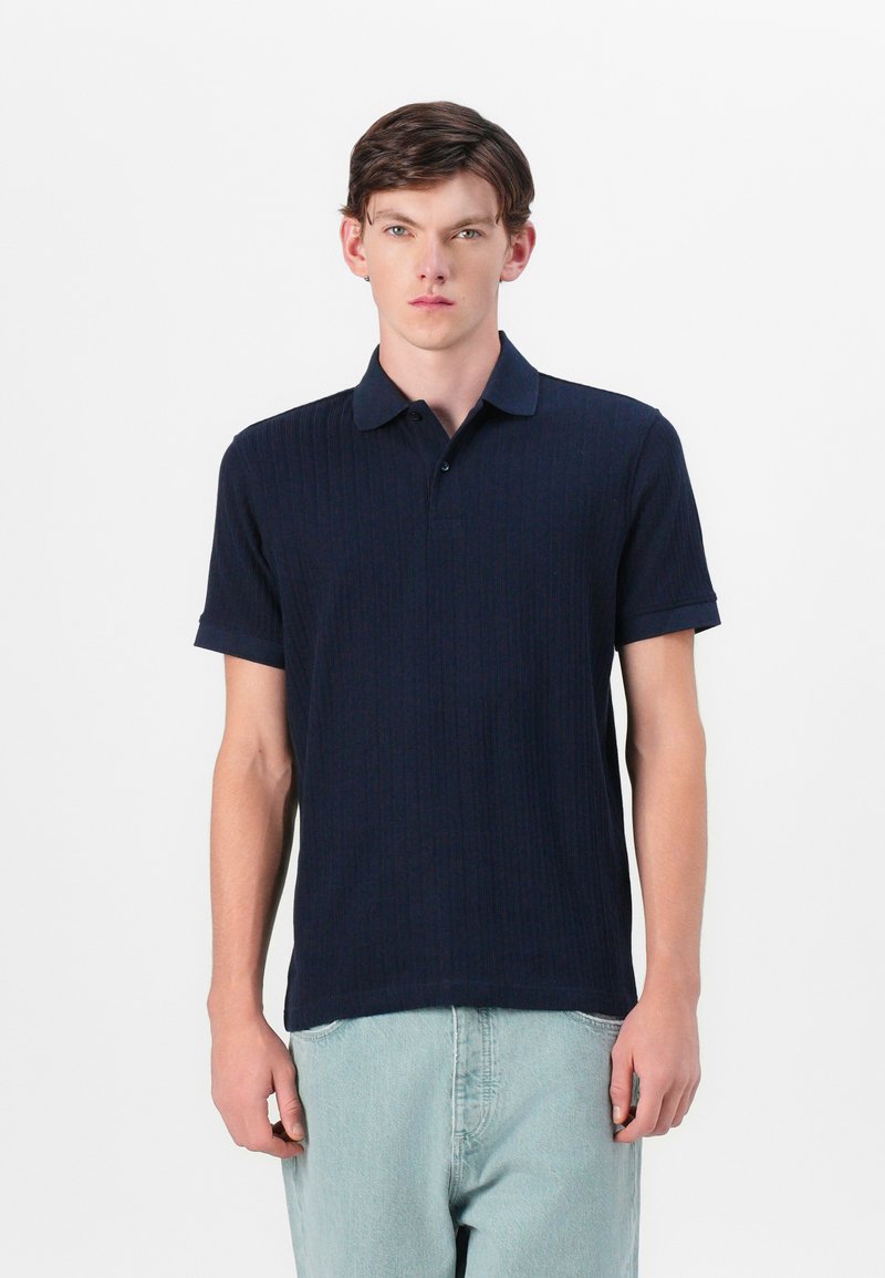 Camisa polo azul marinho com textura ribada, mangas curtas e colarinho clássico. Apresenta uma bainha reta e uma pala com três botões.