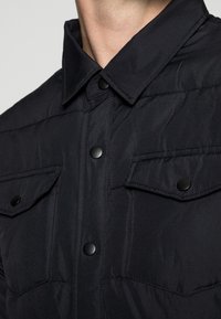 Närbild av en person som bär en svart vadderad jacka med krage och två knäppta bröstfickor.