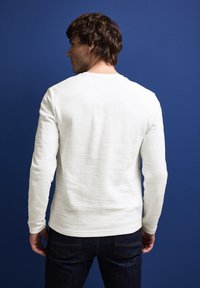 Camisa de manga larga gris claro con una tela texturizada, cuello redondo y mangas con puños, combinada con jeans de mezclilla oscura sobre un fondo azul.
