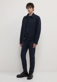 Giacca blu con colletto, chiusura a zip e maniche lunghe, abbinata a pantaloni coordinati e scarpe nere, in piedi contro uno sfondo chiaro.