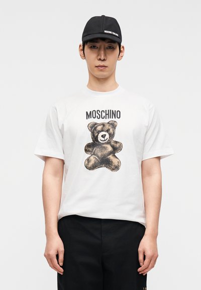 Λευκό βαμβακερό μπλουζάκι με γραφικό αρκούδας και το κείμενο "MOSCHINO" σε μαύρο. Συνδυάζεται με μαύρο καπέλο που φέρει το όνομα της μάρκας.