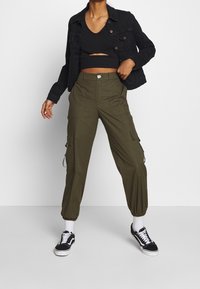 Pantalons cargo vert olive avec des poignets élastiques, plusieurs poches et une fermeture à bouton à l'avant, associés à un haut court noir et une veste en denim.