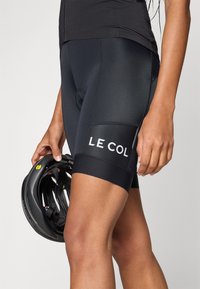 Sorte sykkeltights laget av glatt, elastisk stoff har en fremtredende hvit "LE COL"-logo på låret. En svart hjelm holdes i hånden.