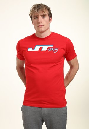 Červené tričko s krátkým rukávem vyrobené z bavlny, na přední straně se modro-bílým logem "JT Racing". Hladká textura, klasický střih.