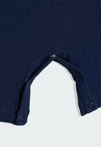 Tmavomodrý rebrovaný overal s viditeľnými patentkami na spodnej strane. Má jemnú textúru a priliehavý strih.