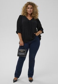 Sort, lette bluse med V-udskæring, bløde ærmer, parret med mørkeblå jeans og en lille sort håndtaske med gyldne detaljer.