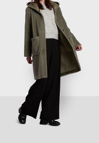 Cappotto con cappuccio verde oliva in un tessuto morbido e testurizzato, con tasche frontali e chiusure a bottoni, abbinato a un maglione grigio e pantaloni larghi neri.