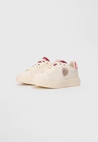 Baskets basses crème avec des accents en velours rose, des lacets crème et un emblème brillant rose et or sur le côté.