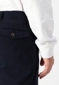 Mornarsko plave kord trousers s bočnom džepom na dugme. Vidljiv bijeli rukav košulje s rebrastim manžetom. Glatka tekstura.