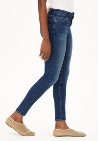 Jean skinny bleu en denim, avec une taille mi-haute, cinq poches et un léger effet délavé. Associé à des chaussures beiges sans lacets.