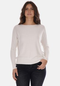 Maglione leggero color crema con scollo ampio, maniche lunghe e polsini a coste, abbinato a jeans blu scuro.
