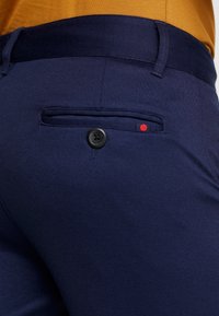 Námořnické modré kalhoty z hladké tkaniny, se zadní kapsou na knoflík a malým červeným akcentem. Pas je tmavší modrý.