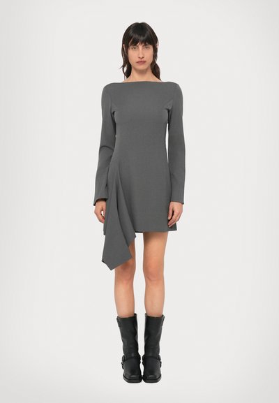 Robe grise à manches longues avec un ourlet asymétrique, réalisée en tissu texturé. Associée à des bottes noires montantes à boucles.
