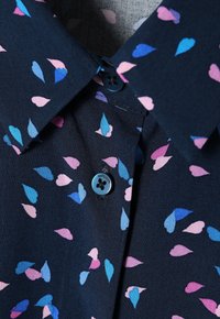 Chemise bleu foncé avec col plié, ornée de formes en pétales rose, bleu et violet, et boutons noirs, dont un gravé du nom de la marque.