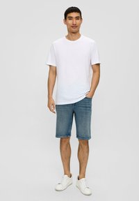 T-shirt en coton blanc, shorts en denim bleu clair et baskets blanches. Le modèle se tient les mains dans les poches, avec des cheveux courts et foncés, sur un fond neutre.