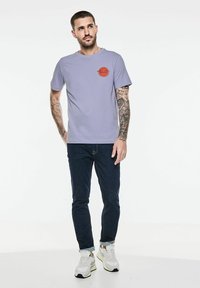 T-shirt en coton violet clair à manches courtes, avec un imprimé graphique rond rouge sur la poitrine, associé à un jean foncé et des baskets blanches.