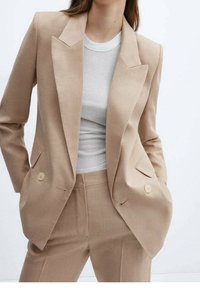 Blazer beige ajusté avec des revers affûtés et des boutons doubles, associé à un t-shirt en jersey blanc. Tissu lisse et silhouette structurée.