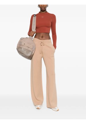 Donna con crop top a maniche lunghe color ruggine e pantaloni beige a gamba larga che tiene una borsa da palestra beige con tracolla.