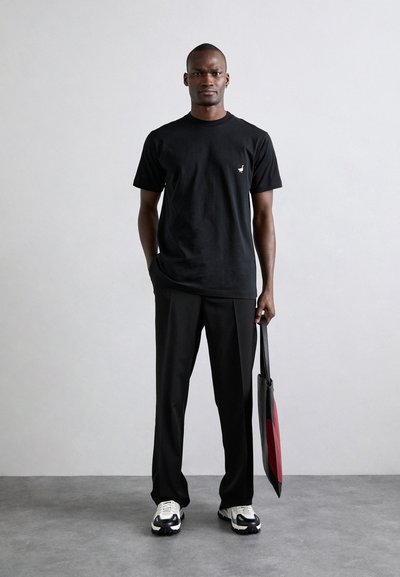 Must värvi puuvillane T-särk lühikeste varrukatega ja väike kuldne logo vasakul rinna piirkonnas, koos mustade pükste ja mustade tossudega, hoides kotti.