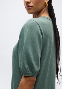 Žena s dlhými zapletenými vlasmi, ktorá nosí zlatý náušník tvaru slzy a zelený pletený top s krátkymi nadýchanými rukávmi.