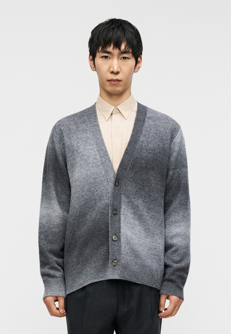 Γκρι καρδιοκάλυμμα με διαβαθμισμένο χρωματισμό  σε απαλό πλεκτό ύφασμα, με λαιμόκοψη σε σχήμα V, κλείσιμο με κουμπιά και μακριά μανίκια. Φορεμένο πάνω από ανοιχτό μπεζ πουκάμισο.