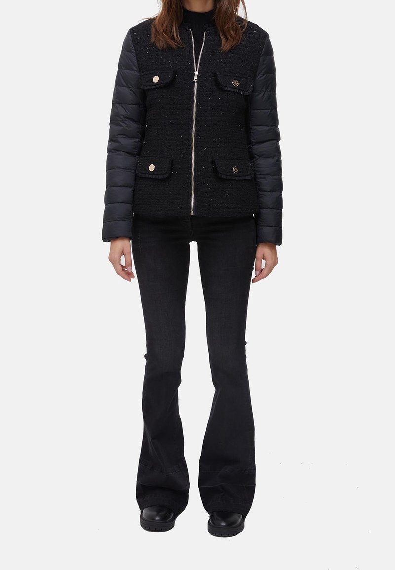 Femme portant une veste noire zippée avec des manches matelassées et des poches à rabat, associée à un pantalon noir évasé et des chaussures noires sur fond blanc.
