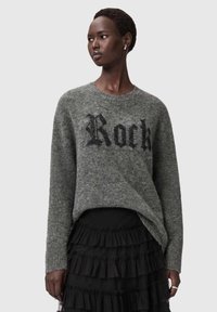 Nainen pukeutuneena harmaaseen pörröiseen neulepuseroon, jossa lukee "Rock", ja mustaan kerrokselliseen röyhelöhameeseen, katsomassa vasemmalle harmaalla taustalla.