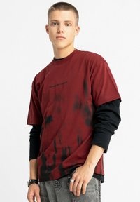 Czerwona i czarna koszulka tie-dye z krótkimi rękawami, z okrągłym dekoltem i nadrukiem graficznym na przodzie. Nosi się ją na czarnej koszuli z długim rękawem.