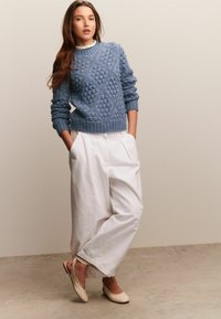 Mujer vestida con un suéter de punto texturizado azul, pantalones blancos de pierna ancha y unos zapatos de tacón abierto beige, de pie con las manos en los bolsillos, frente a un fondo liso.
