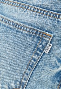 Vaaleansininen farkkukangas lähikuvassa, jossa näkyy takatasku oranssilla ompelella ja pieni valkoinen etiketti sauman kohdalla.