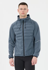 Ung mand iført en blå hættejakke med lynl�ås og polstret front samt sorte leggings, der står foran en hvid baggrund.