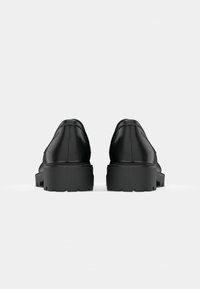 Paire de chaussures habillées en cuir noir à semelle épaisse, vues de dos sur un fond gris clair.