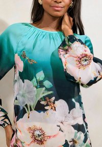 Blusa di seta con sfondo degradé che va dal teal al scuro, caratterizzata da grandi stampe floreali e motivi di farfalle decorate su maniche ampie.